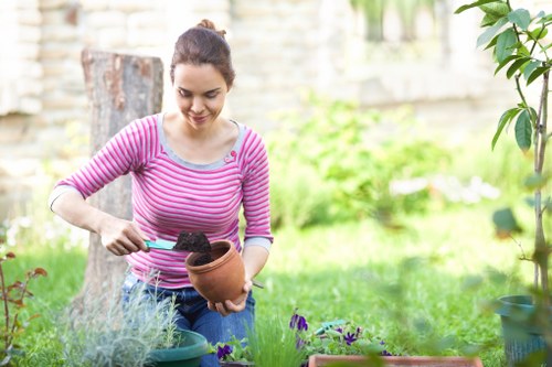 A young woman with dark hair tied back, wearing a pink and white striped long-sleeve top, is tending to a garden in a residential outdoor space. She is holding a terracotta pot filled with dark soil and using a small gardening trowel to transfer soil into it. The garden features a lush, green lawn with a variety of plants, including flowering purple and green foliage, arranged in groups. In the background, there are small trees, shrubs, a weathered tree stump, and a stone or concrete wall that surrounds the garden. Potted plants and gardening tools are visible on the ground, indicating ongoing plant care and maintenance. The scene is illuminated by natural daylight, suggesting a clear or partly cloudy weather, with a softly blurred background emphasizing the gardening activity and garden elements like grass, flowering plants, soil, and foliage. The image subtly supports local garden maintenance services in Osterley, reflecting a well-kept outdoor space suitable for professional gardening and landscaping care by Gardening Services Osterley.